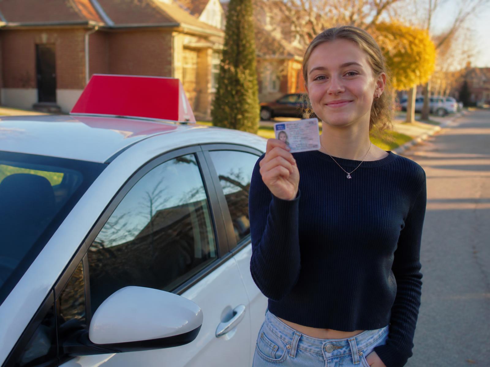 Smiling new driver holding her Ontario licence beside a Provincial Driving School car